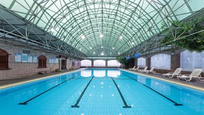 Ein Hallenbad mit Glasdach, deutlich abgetrennten Bahnen und Liegestühlen entlang der Seiten.