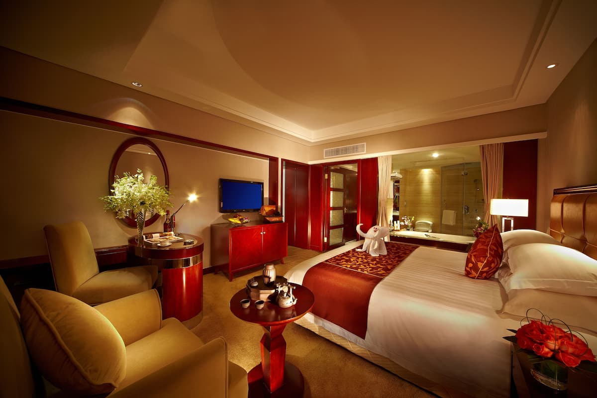 Habitación de hotel con una cama grande, un área de estar con una mesa y sillas, un televisor y un baño visible a través de una puerta abierta.