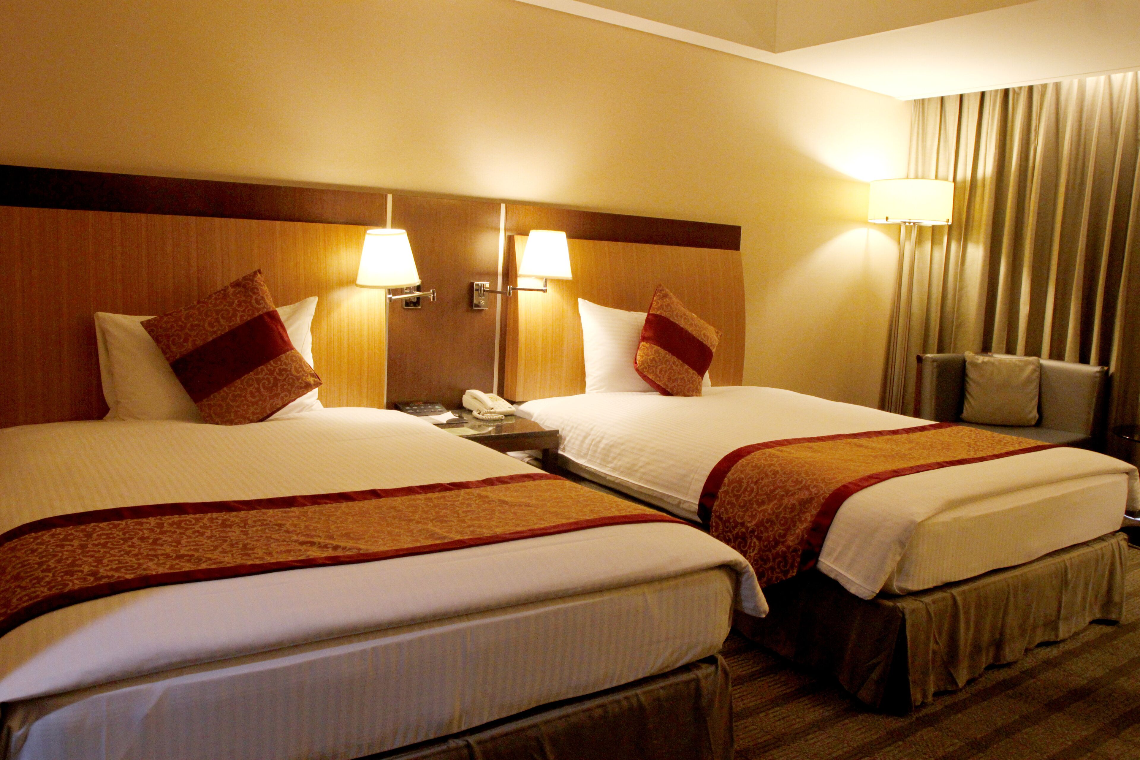 Two adjacent hotel beds with matching headboards, bedside lamps, and a neutral-toned curtain.