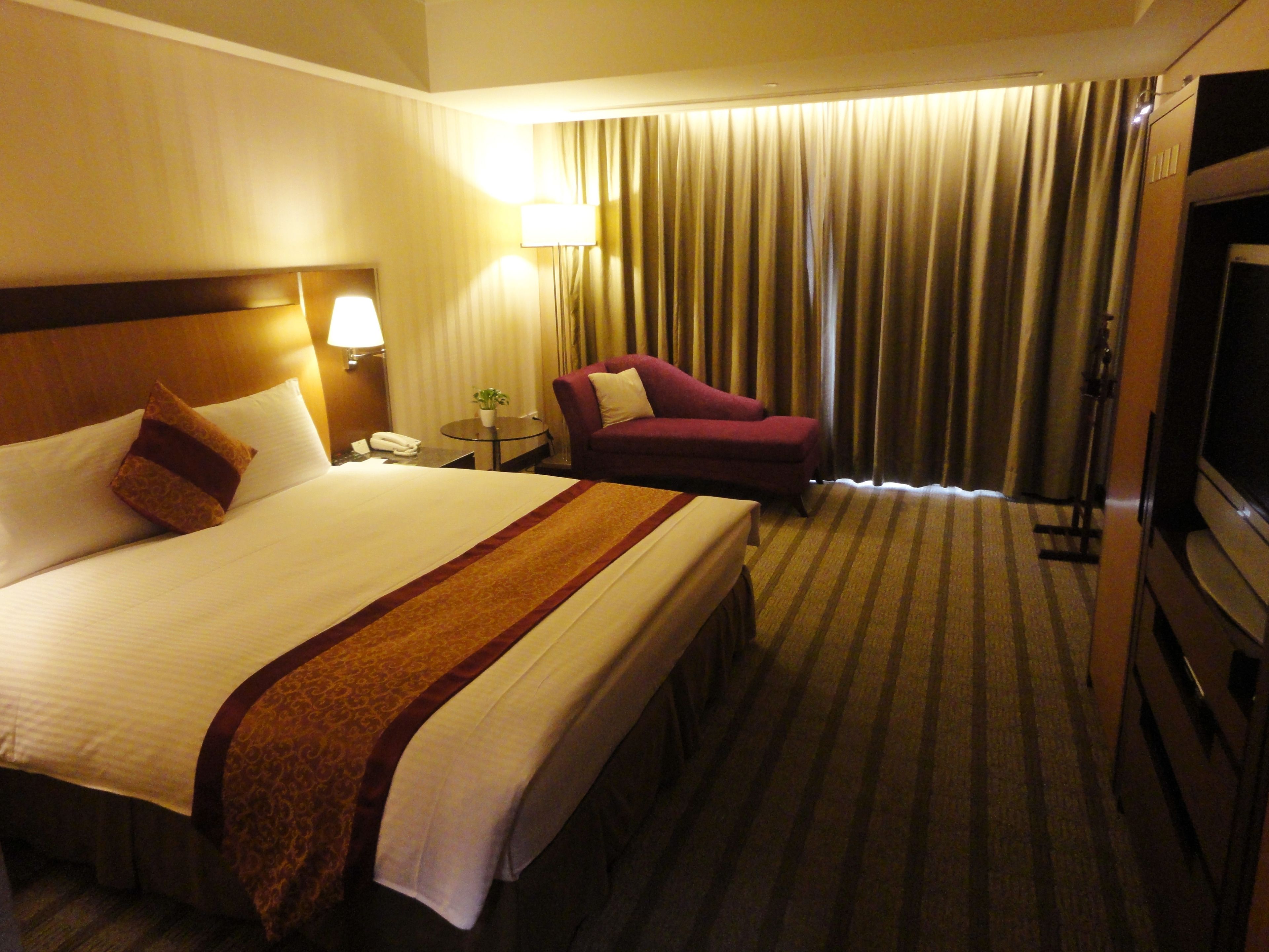 A hotel room with a large bed, a red armchair, a TV, and a lamp.