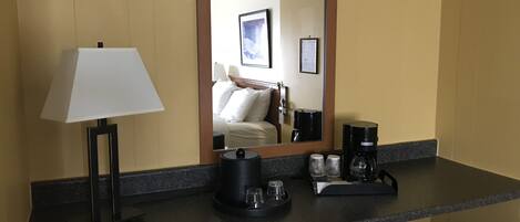 Quarto de hotel com bancada escura, luminária, cafeteira e espelho refletindo uma cama e um quadro emoldurado.