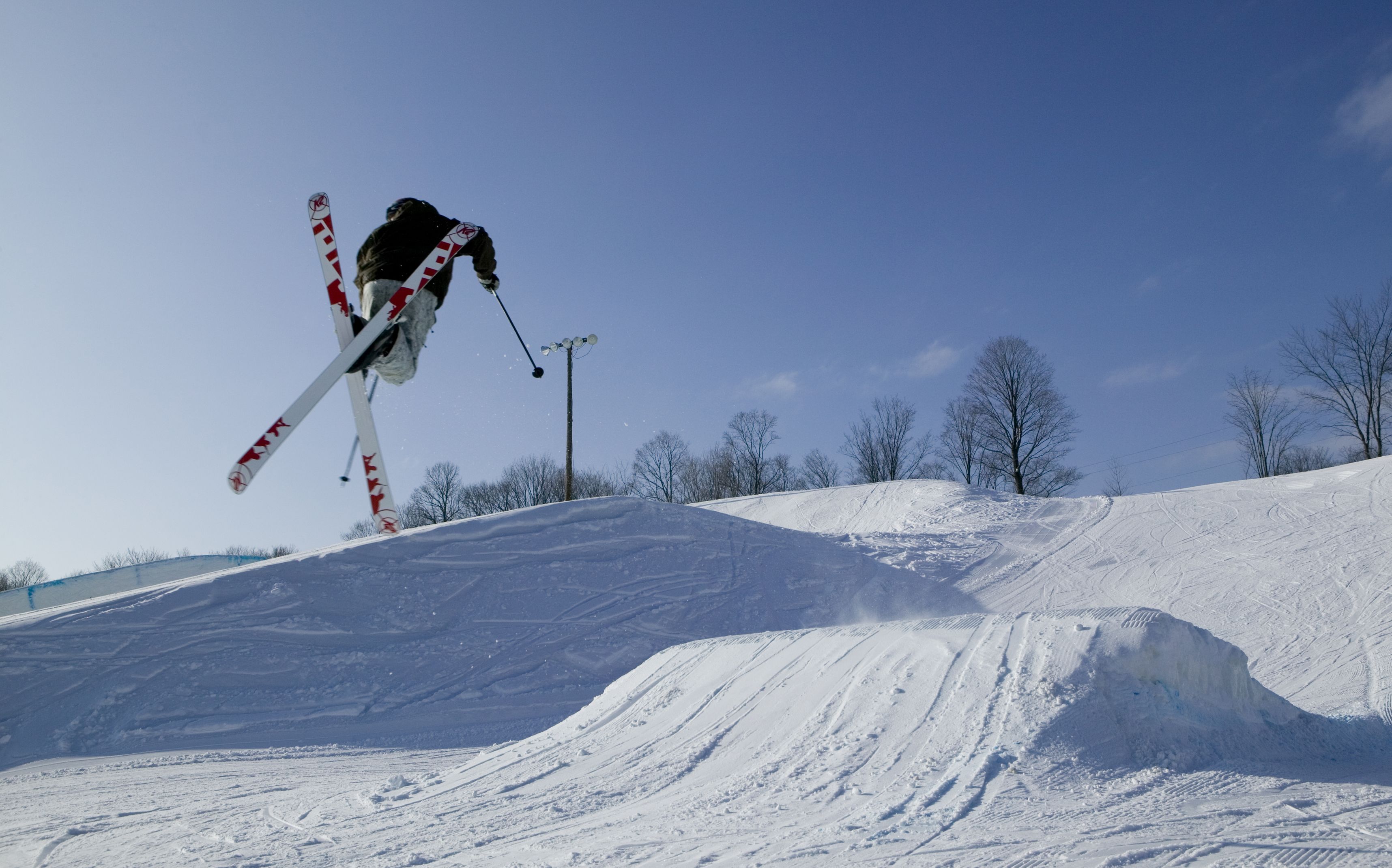 Un tremplin de ski avec un skieur en plein vol, une pente enneigée et des arbres sans feuilles en arrière-plan.
