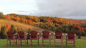 Four Adirondack chairs facing a scenic view of a forested hillside with autumn foliage. - Otsego Resort (Gaylord)