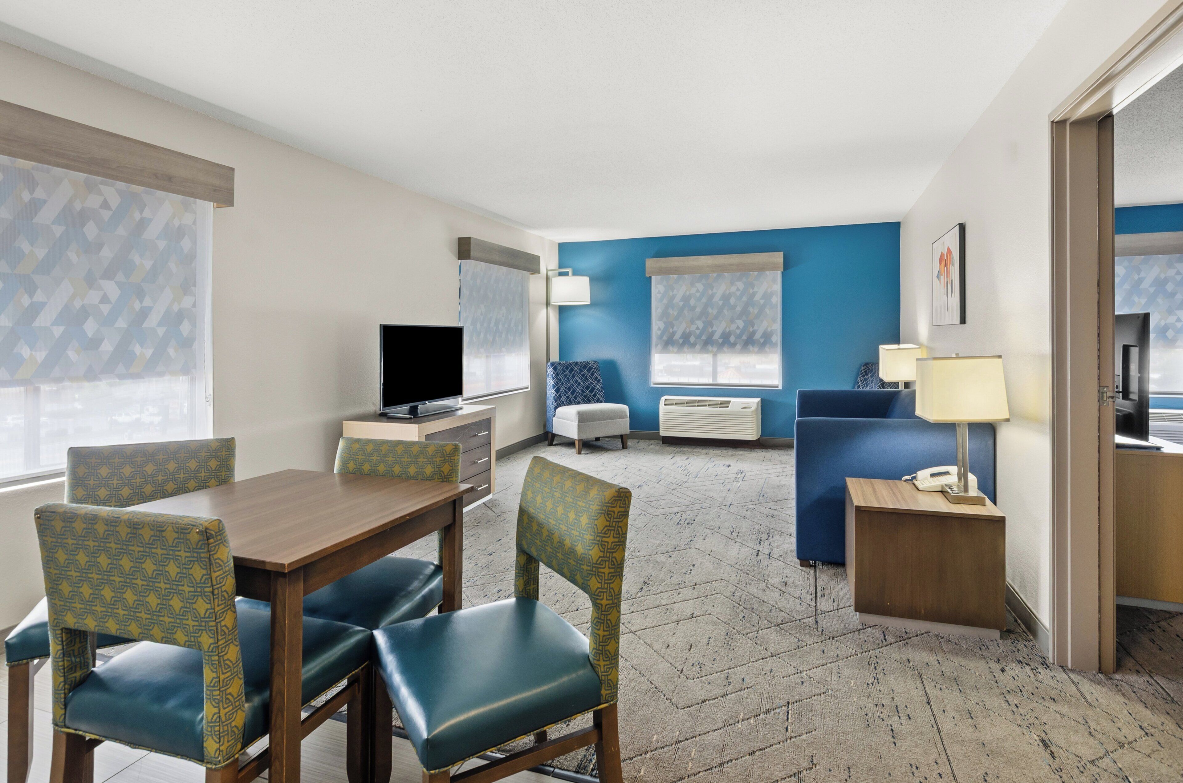 A hotel room with a wooden dining table, blue cushioned chairs, a sofa, a TV, a lamp, and a small bedside table.