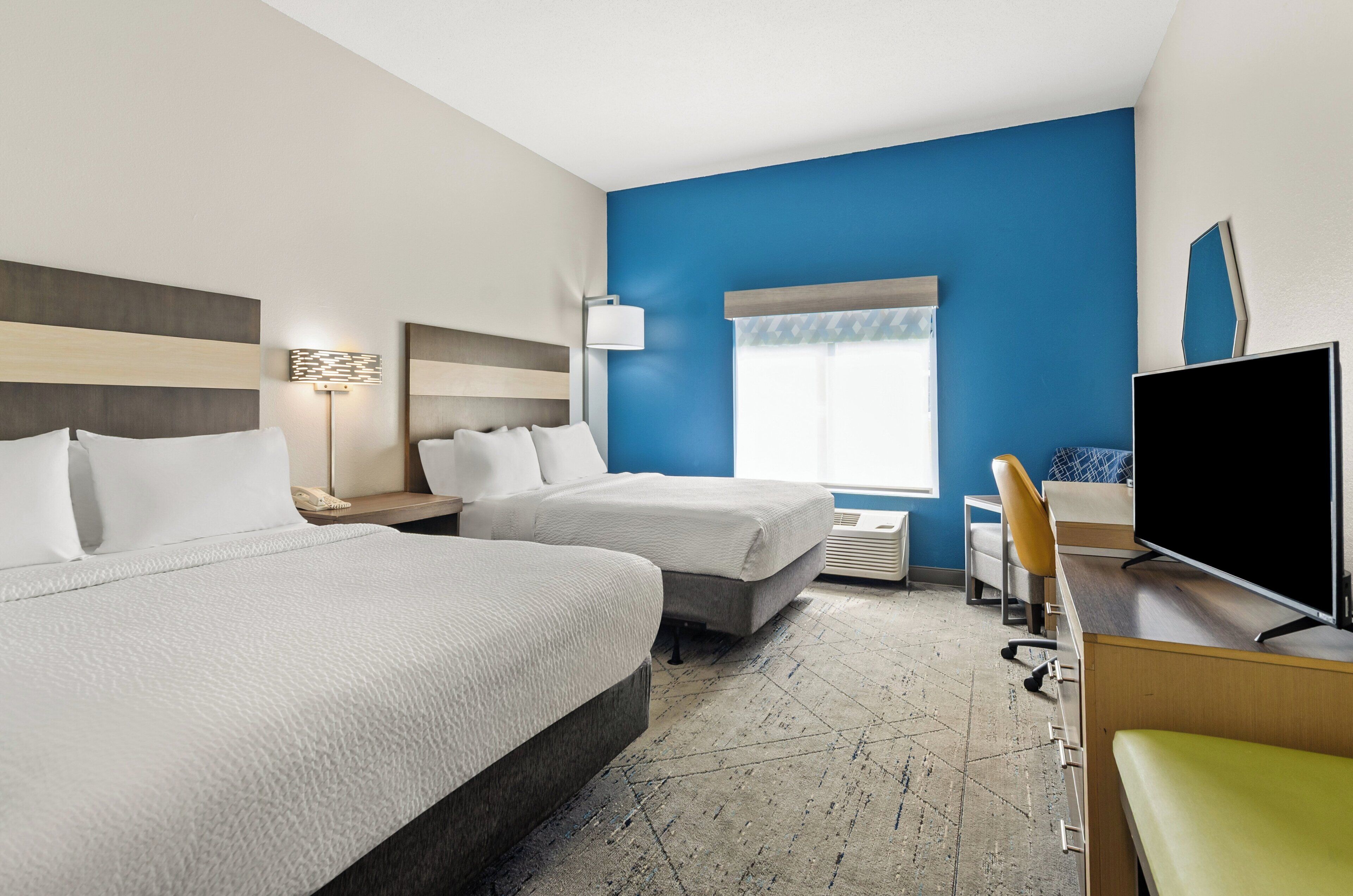 A hotel room with two beds, a desk with a flat-screen TV, a chair, and a window with blue walls and a view.