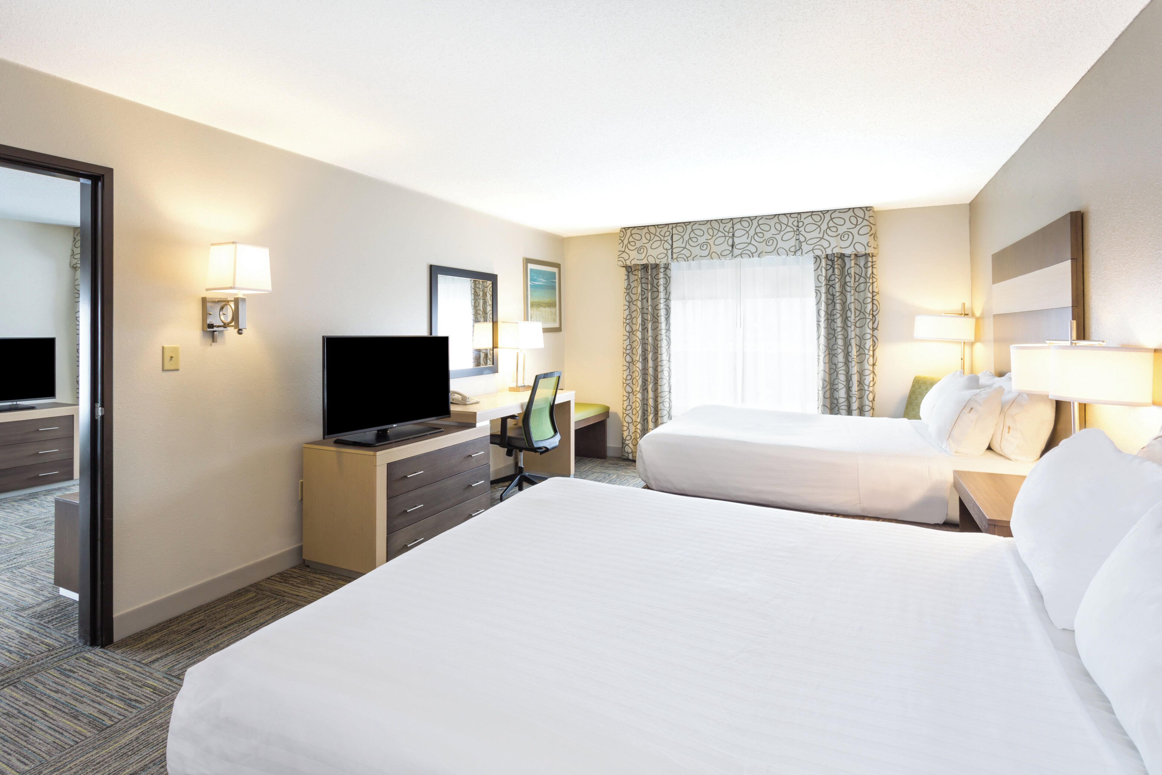 Une chambre d’hôtel avec deux lits, un bureau avec une télévision, une chaise et une fenêtre avec des rideaux.