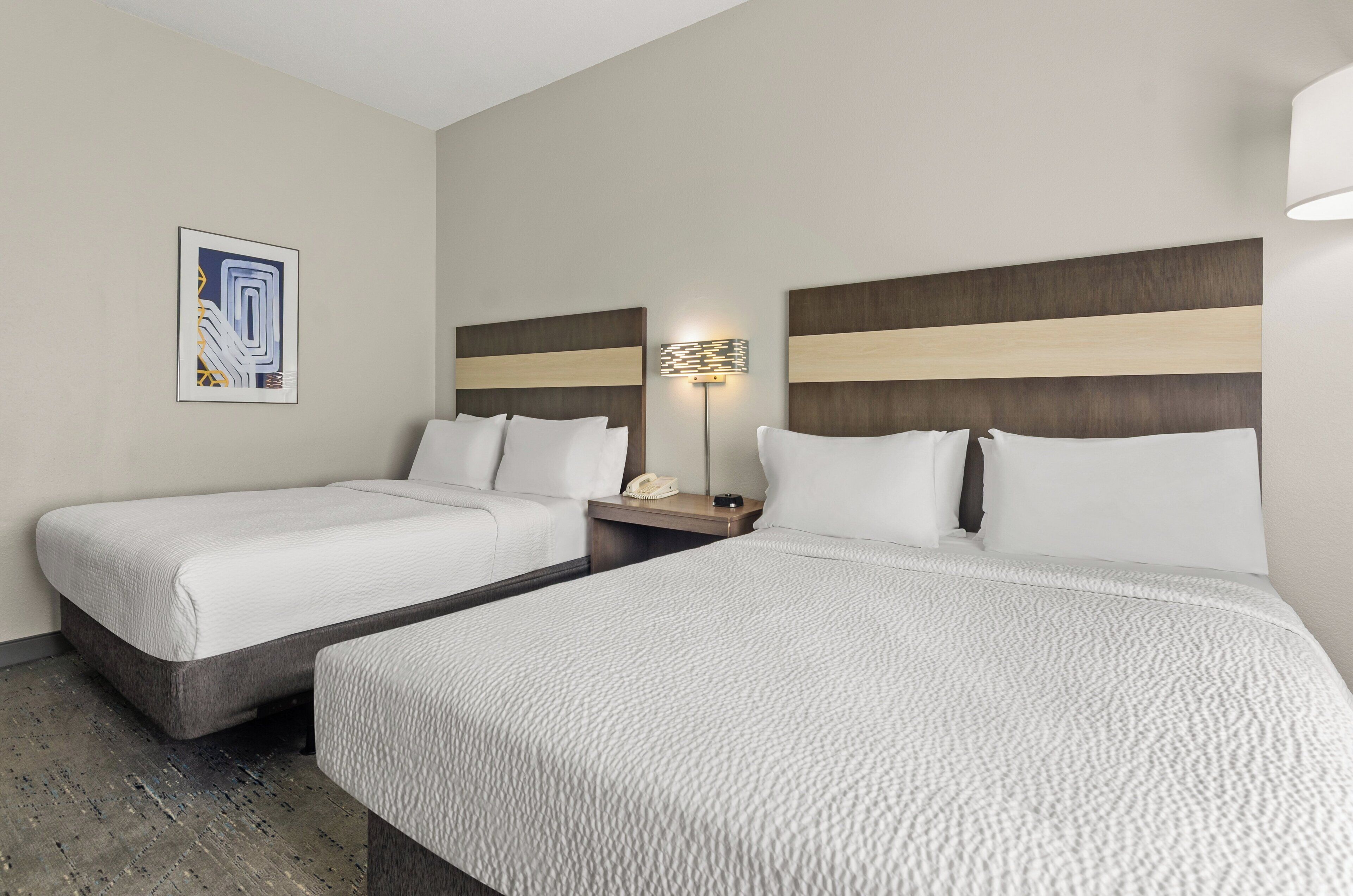 A hotel room with two beds, a bedside table with a lamp, and a framed picture on the wall.