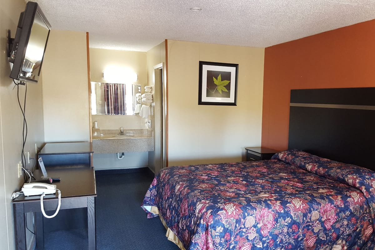 Ein Hotelzimmer mit Bett, Schreibtisch mit Telefon, Spiegel, Waschbecken und einem gerahmten Bild an der Wand.