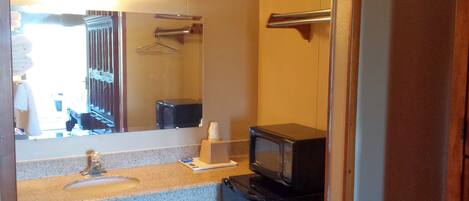 A compact hotel bathroom with a sink, mirror, towel rack, and a small refrigerator.