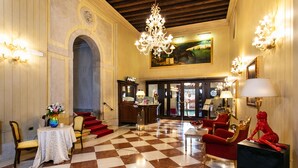 A lobby with a checkered floor, red and gold chairs, a chandelier, and a painting on the wall. - Ca' Vendramin Zago (Venice)