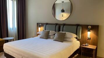Una habitación de hotel con una cama grande, un espejo redondo en la pared, una mesita de noche con una lámpara y una pequeña mesa redonda.