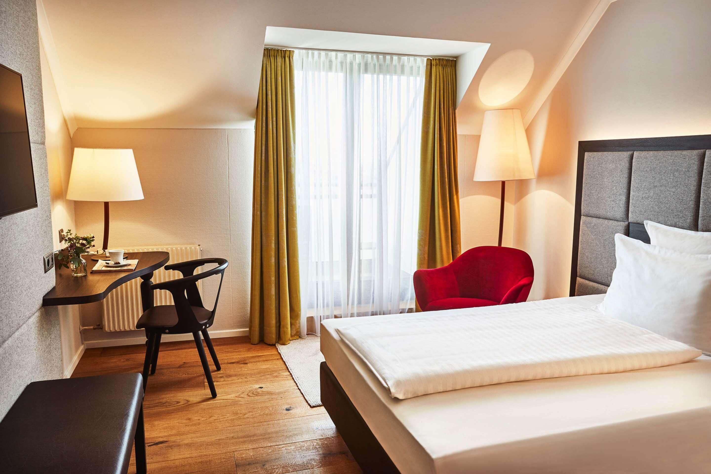 Habitación de hotel con cama, escritorio y silla, una silla roja, dos lámparas y una ventana con cortinas.