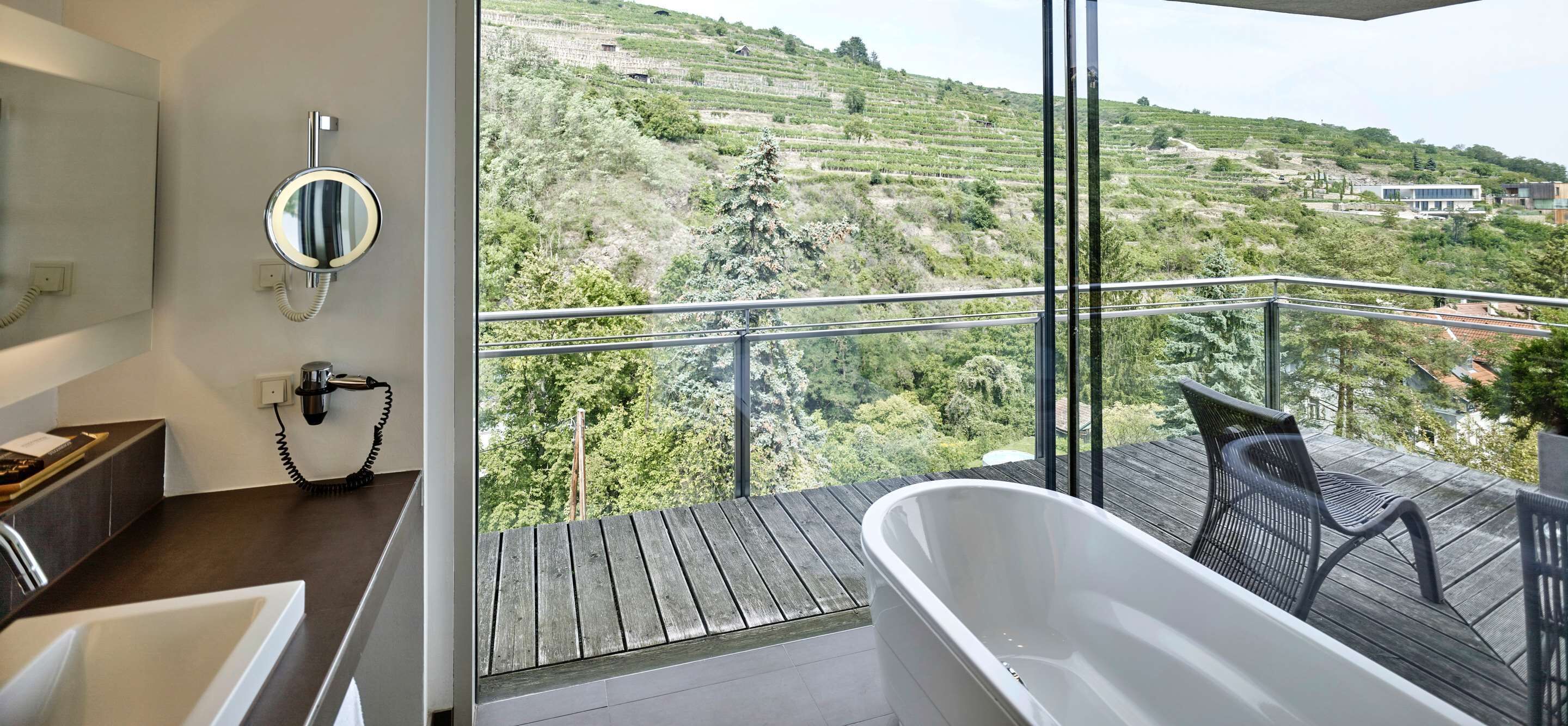 Un baño con una bañera grande, un espejo y un secador de pelo. El baño tiene piso de madera y un lavamanos. El balcón cuenta con piso de madera y vistas a un viñedo y un edificio a lo lejos.