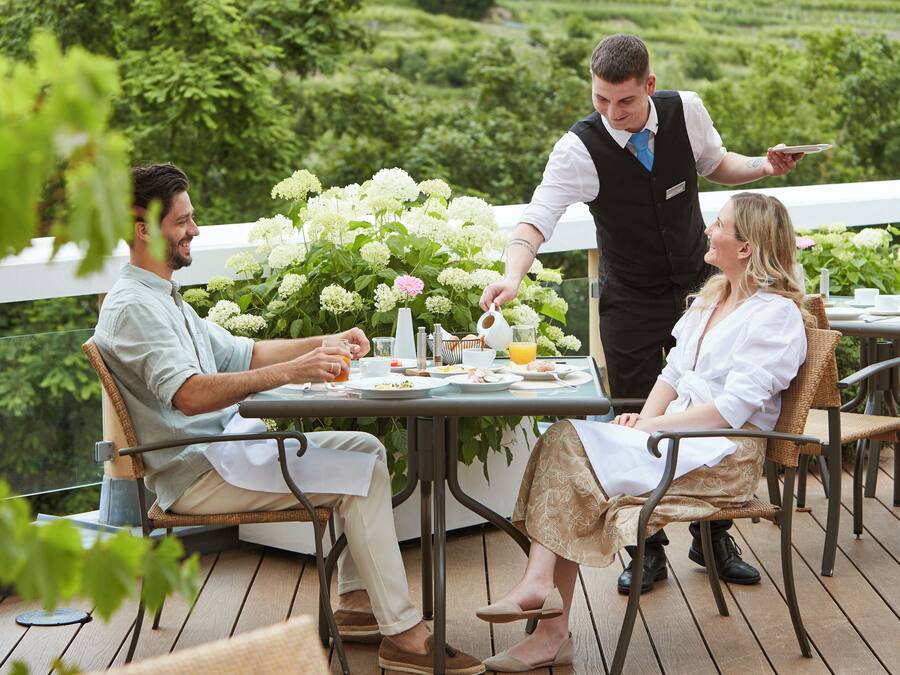 Eine Terrasse mit Tisch für drei Personen, umgeben von Grünpflanzen und einem weißen Geländer.