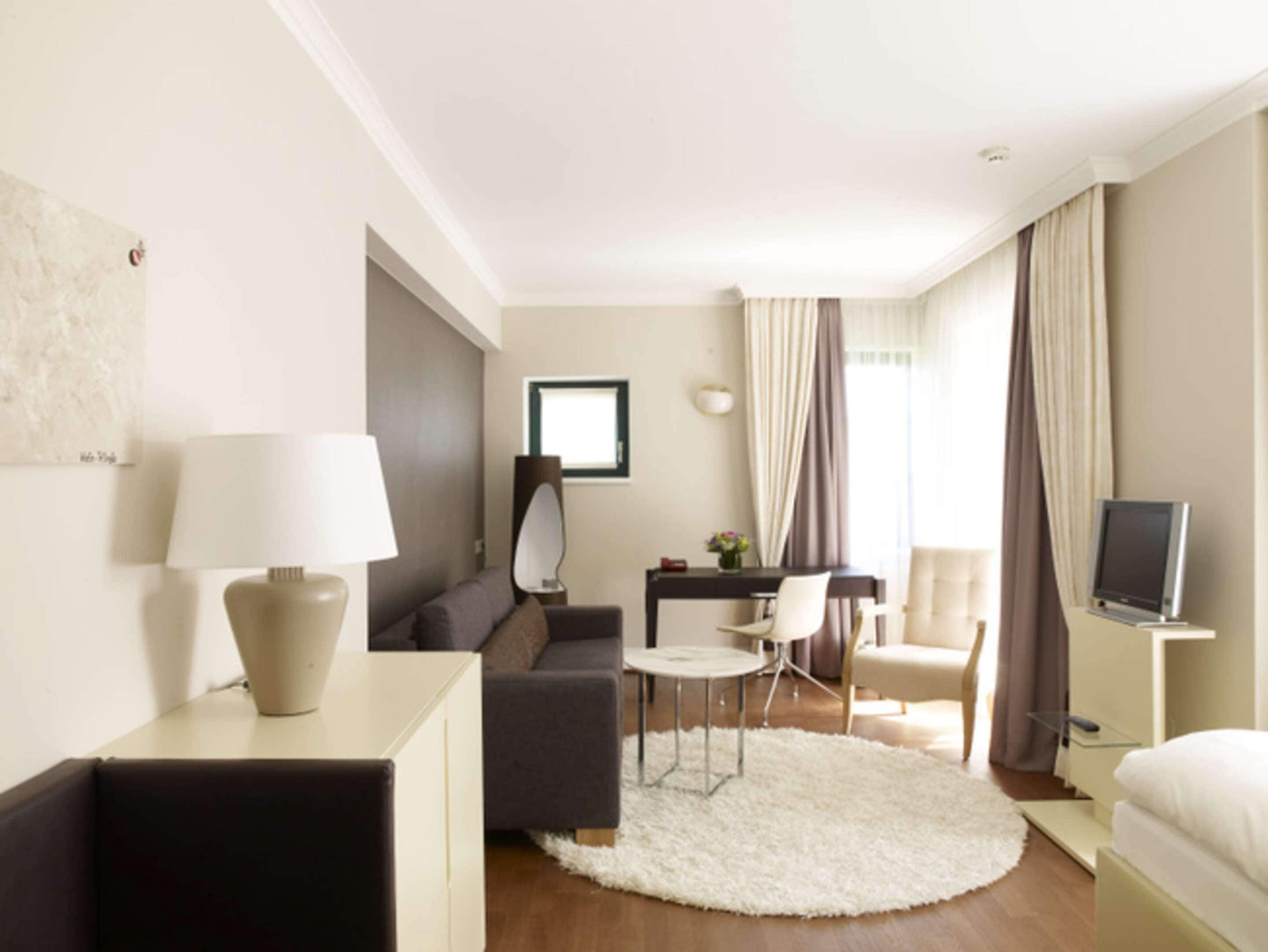 Una habitación de hotel moderna con sofá, mesa de comedor, una silla, televisor y un ventanal con cortinas.