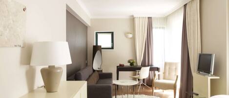Una habitación de hotel moderna con sofá, mesa de comedor, una silla, televisor y un ventanal con cortinas.