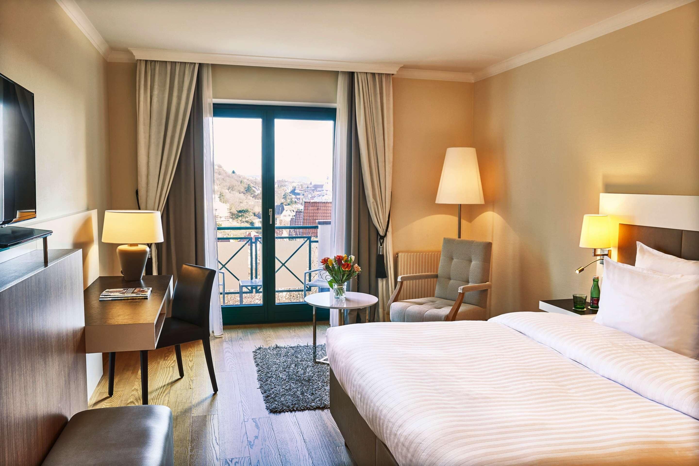 Habitación de hotel con cama, escritorio, silla, dos lámparas, una pequeña mesa con flores y vistas a través de un ventanal grande.