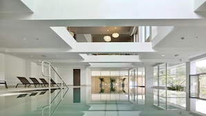 Una piscina interior moderna con paredes de vidrio transparente, una escalera central e iluminación contemporánea.