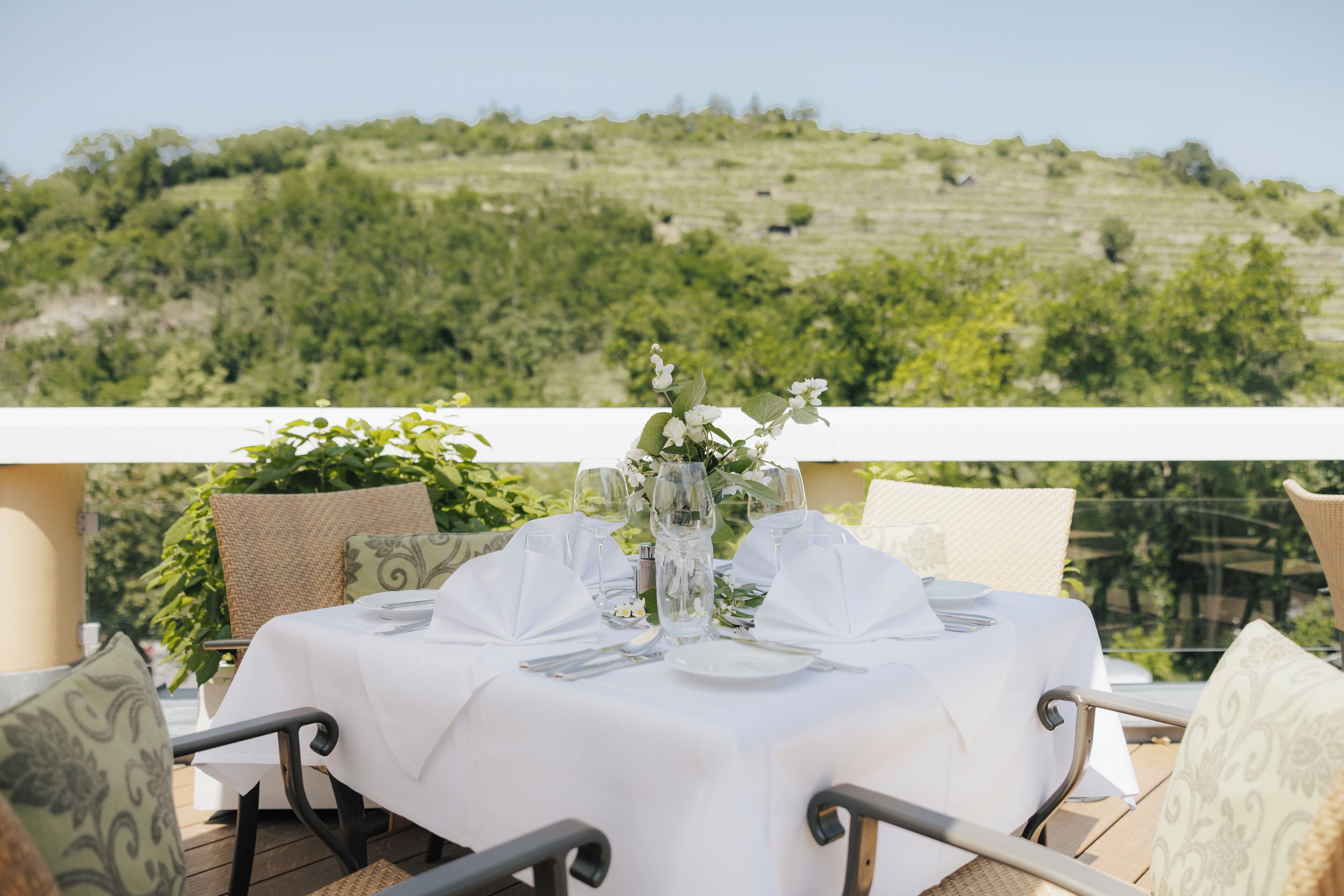A table set for two with a white tablecloth, two chairs, and a vase with flowers. The table is on an outdoor terrace with a view of a green hillside.