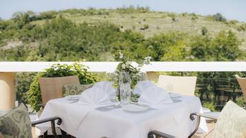 A table set for two with a white tablecloth, two chairs, and a vase with flowers. The table is on an outdoor terrace with a view of a green hillside.