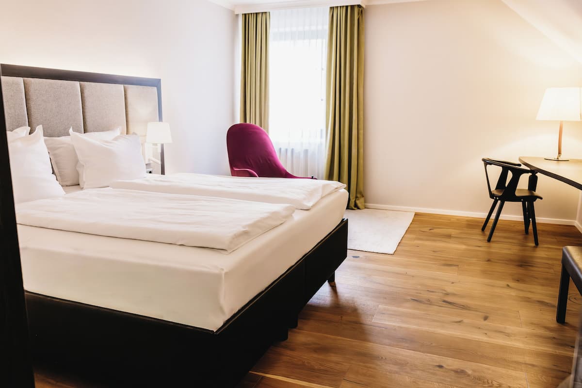 Habitación de hotel con cama doble, escritorio con silla, una silla roja y ventana con cortinas verdes.
