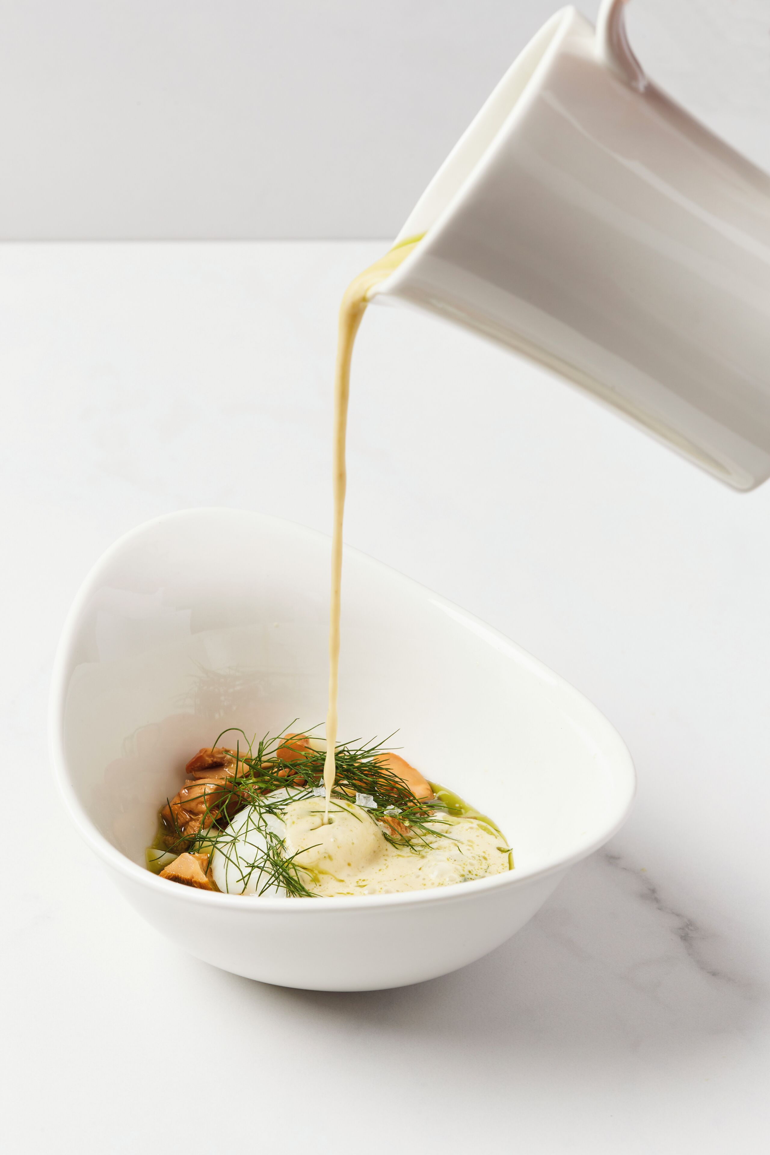 A white bowl with a creamy liquid being poured into it, containing a food item with herbs.