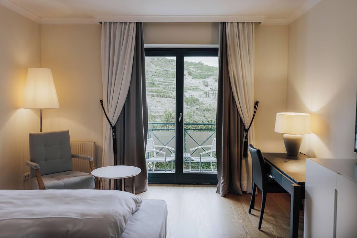 Habitación de hotel con cama, dos sillas, escritorio, mesa y balcón con cortinas.
