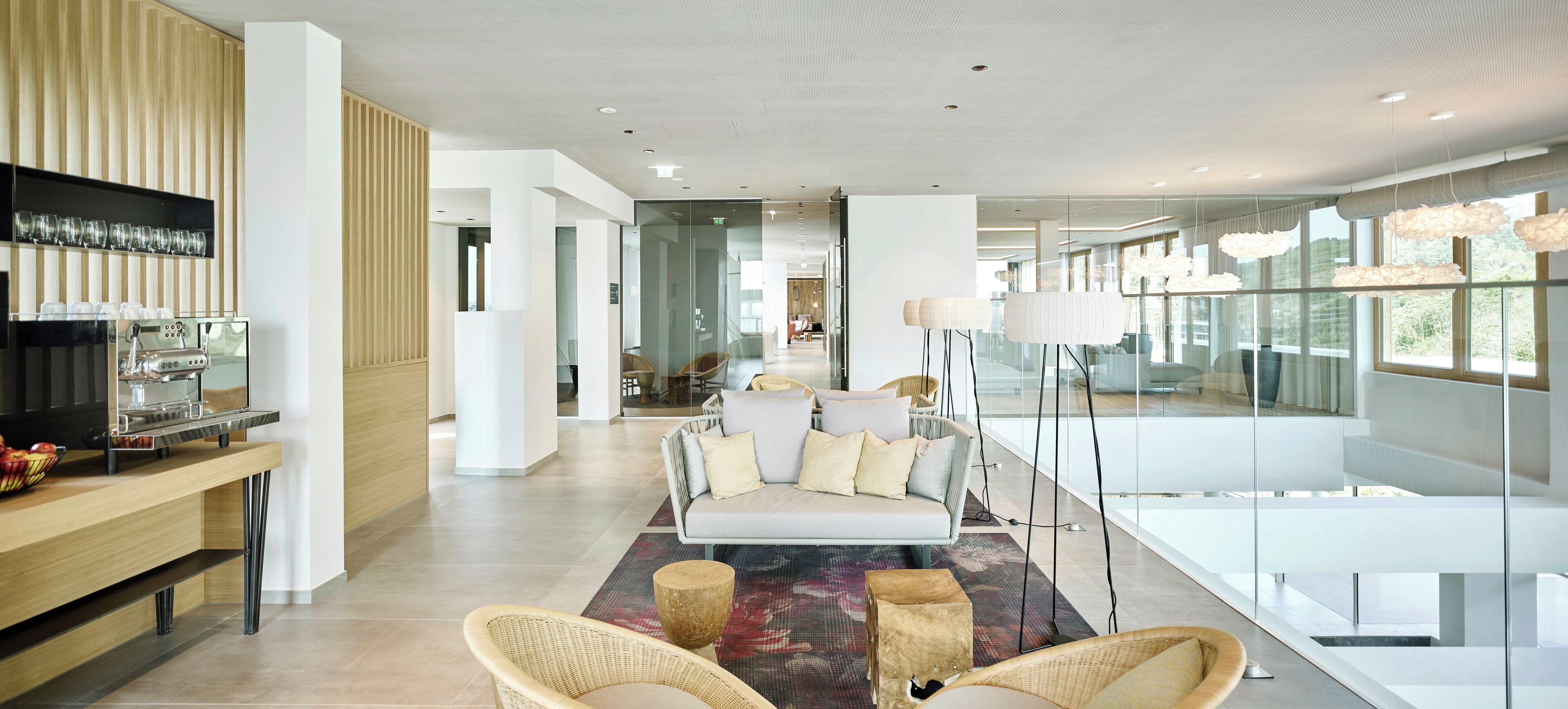 A modern living room with a sofa, wicker chairs, a coffee machine, and a glass partition.