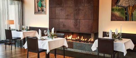 Un comedor con mesas preparadas para dos, una chimenea encendida y obras de arte en las paredes.