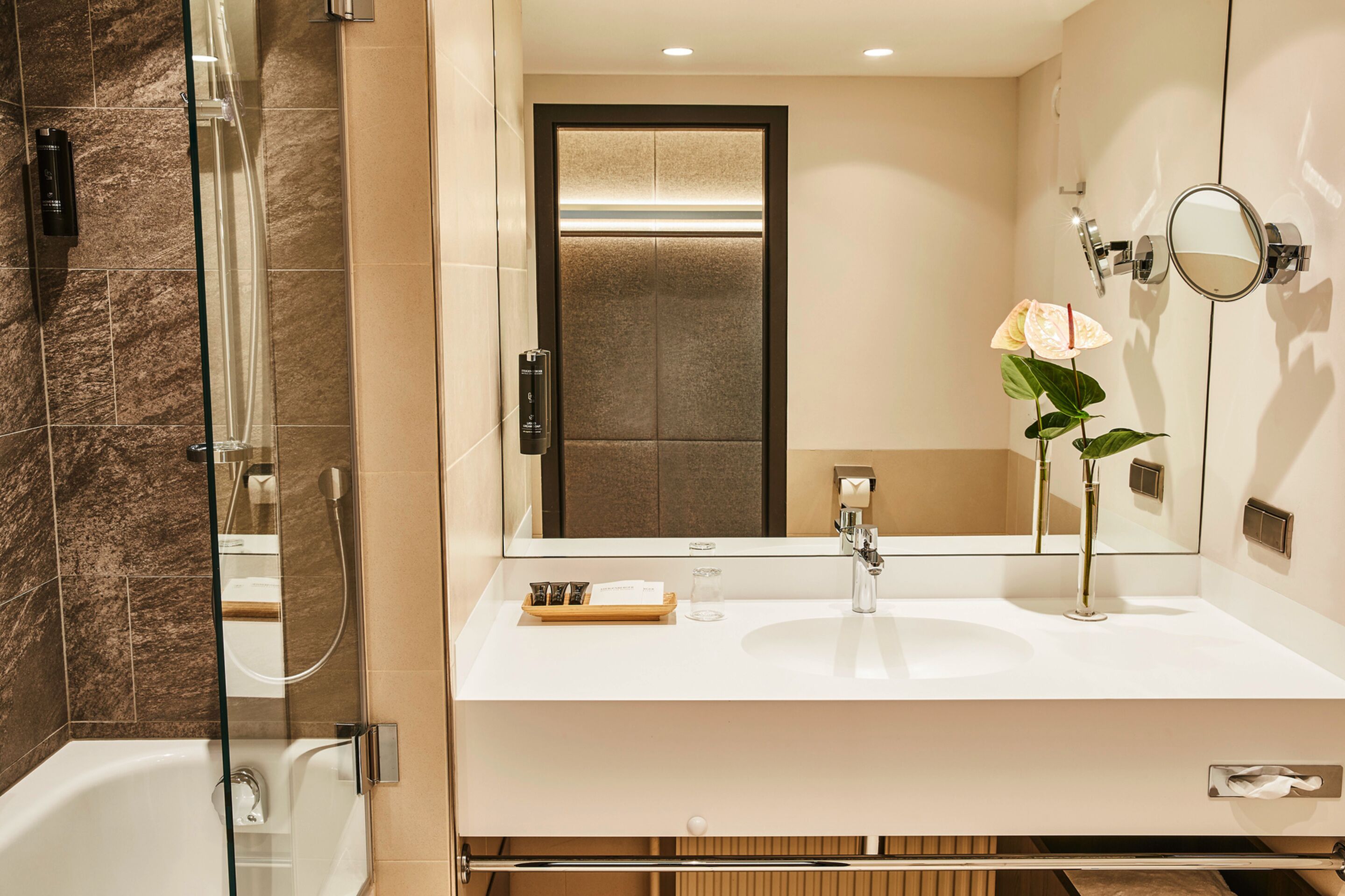 Un baño moderno con ducha de vidrio, un lavamanos blanco con espejo y un florero con una sola flor.