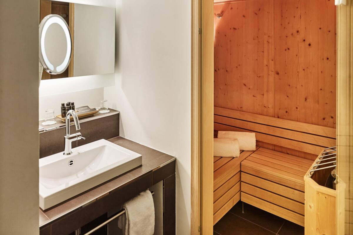 Un baño moderno con lavamanos, espejo y una sauna de madera visible a través de una puerta abierta.