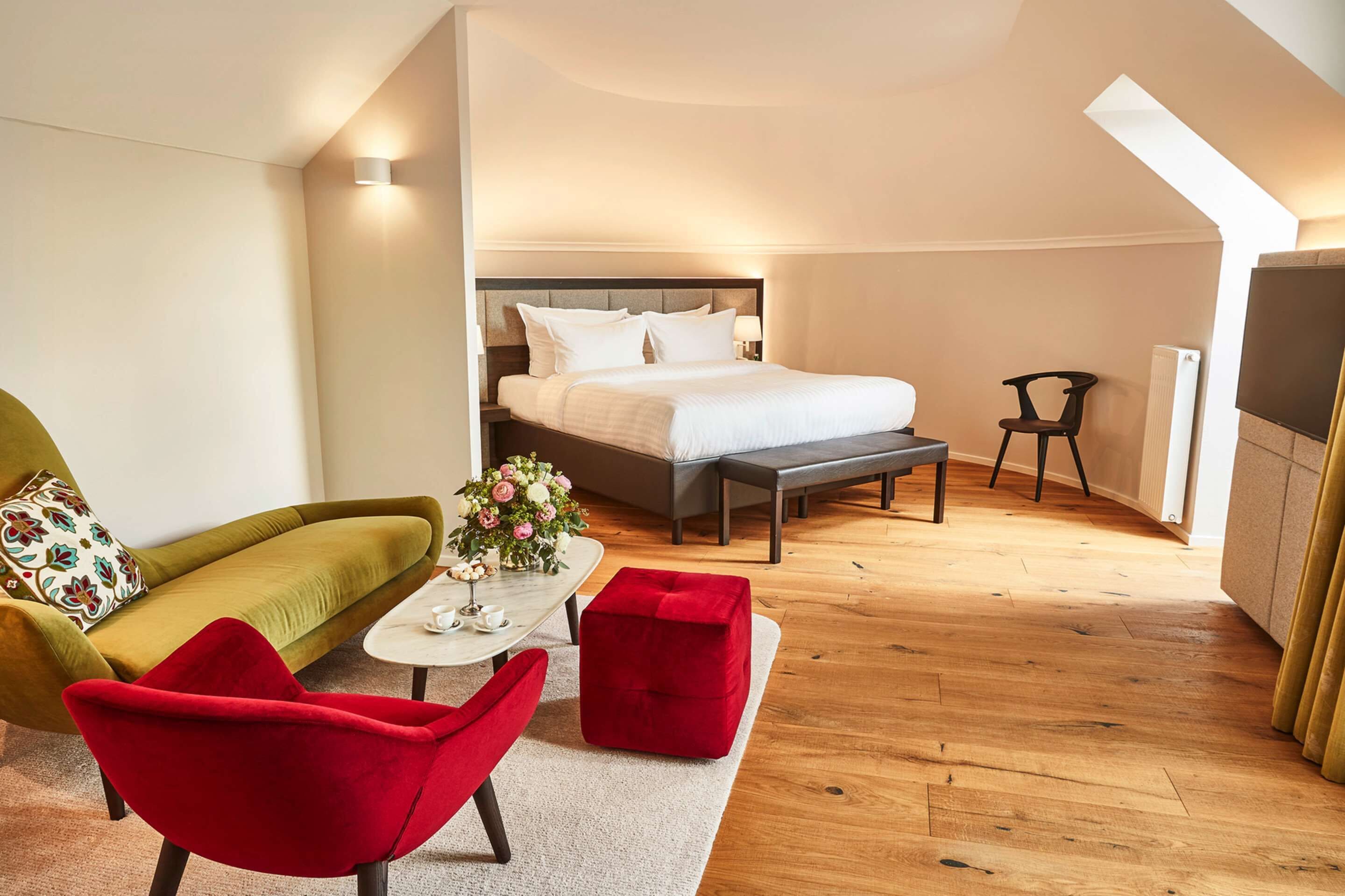 A modern hotel room with a large bed, a green sofa, a red armchair, and a small table with a vase of flowers.