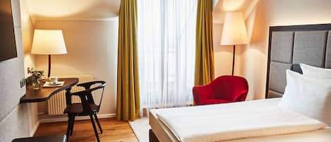 Una habitación de hotel con cama, escritorio y silla, una silla roja, dos lámparas y una ventana con cortinas.