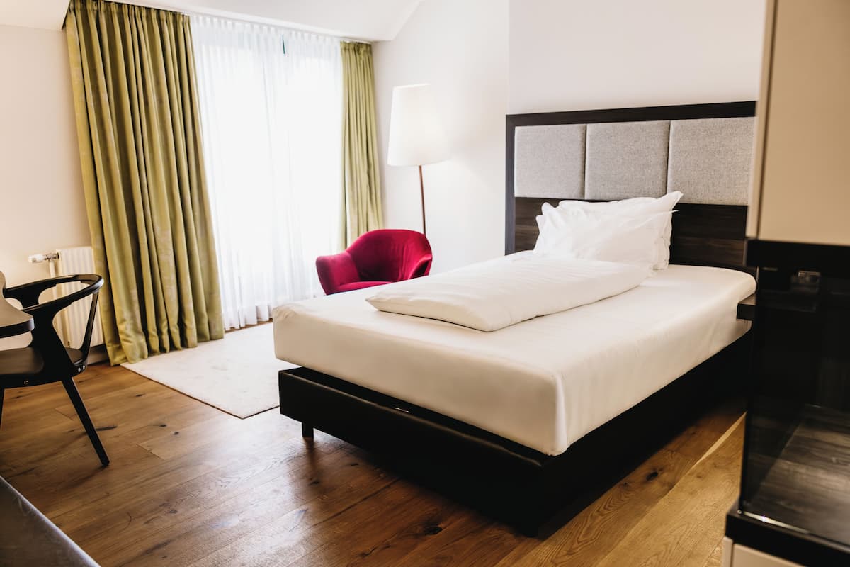 Habitación de hotel con una cama grande, una silla roja, una lámpara y vistas al exterior a través de cortinas.