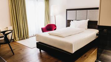 Hotelkamer met een groot bed, een rode stoel, een lamp en uitzicht op het buitenleven door gordijnen.