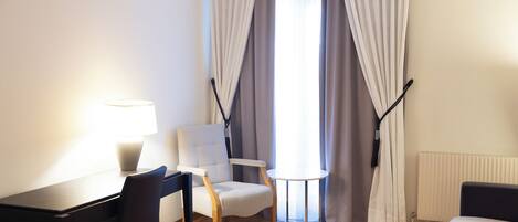 Habitación de hotel con piso de madera, un sofá blanco, una pequeña mesa redonda, un sillón blanco, un escritorio con lámpara y una ventana con cortinas.