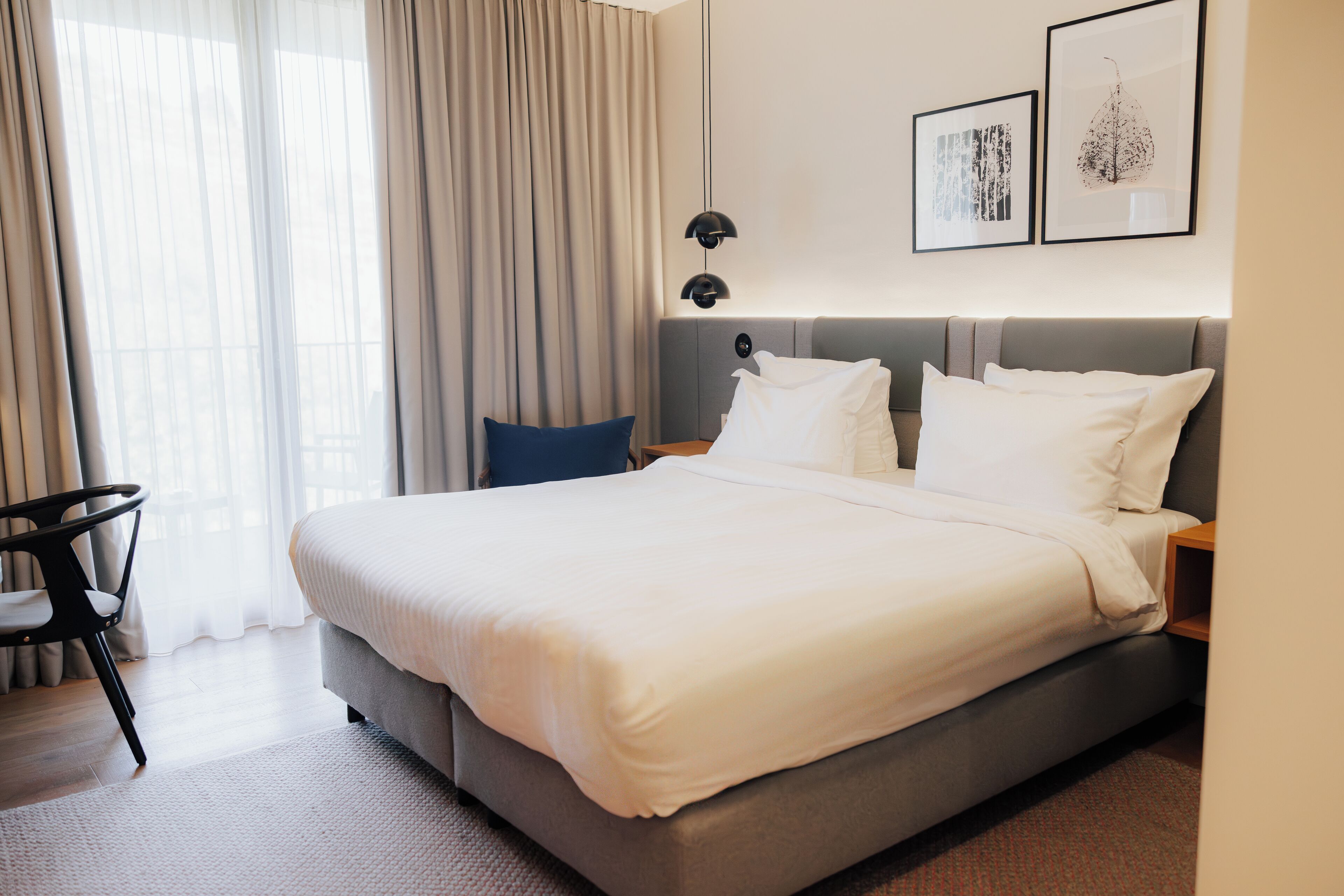 A modern hotel room with a large bed, a bedside table, a chair, and a window with curtains.