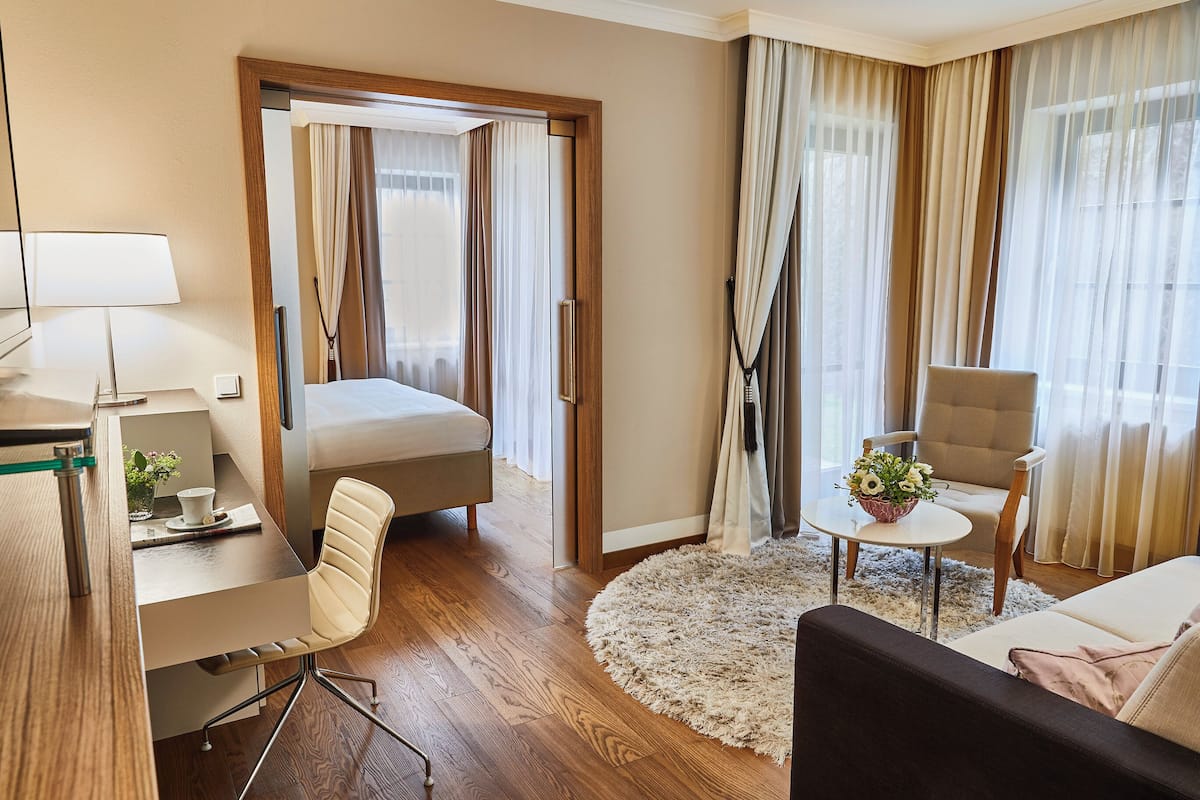 Una habitación de hotel moderna con piso de madera, una cama, un escritorio con lámpara, una silla, un sofá y una pequeña mesa con un florero.