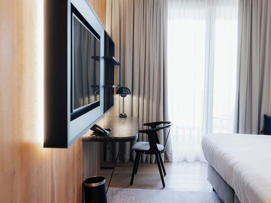 A hotel room with a bed, a desk with a chair, a TV mounted on the wall, and a window with curtains.