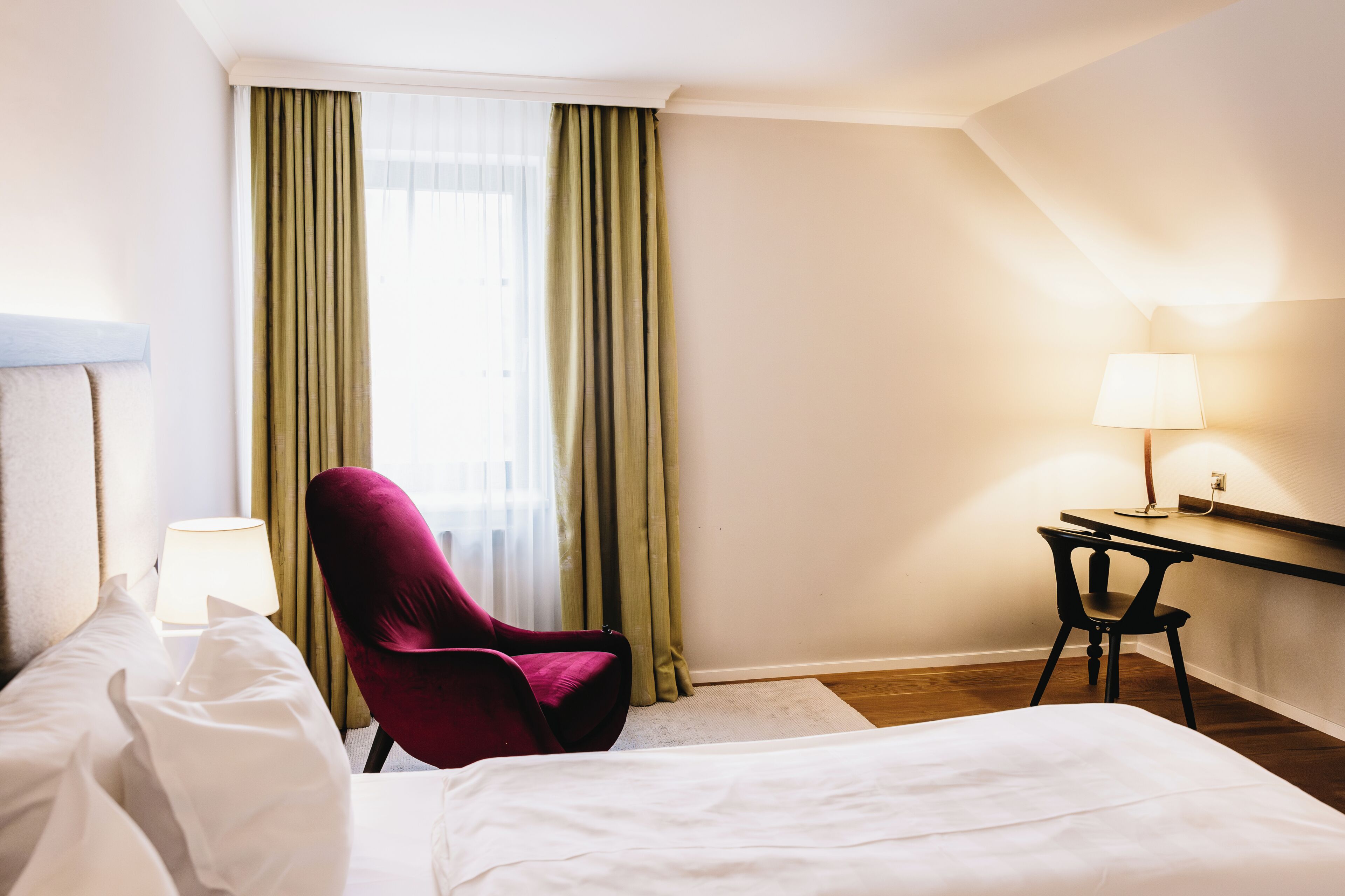 Una habitación de hotel con una cama, una silla roja, un escritorio con lámpara y una ventana con cortinas.