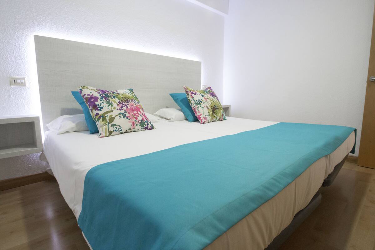 A neatly made bed with a teal bedspread and floral-patterned pillows, a wooden headboard, and a white wall.