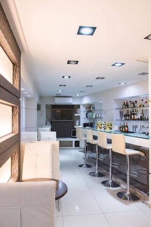A modern bar area with white bar stools, a glass bar counter, and a seating area with white sofas. - Epidavros Hotel (Athens)
