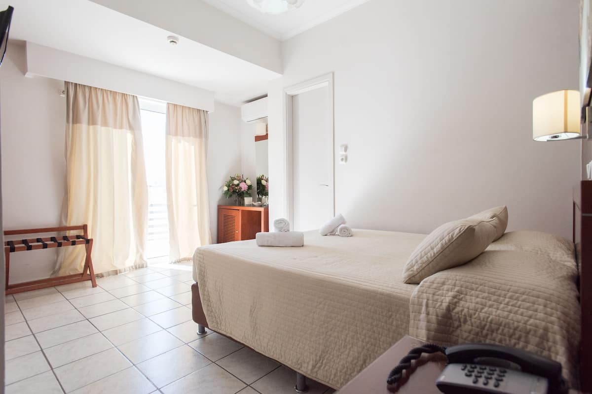 Una habitación de hotel con cama, mesita de noche con teléfono, banco de madera y ventana con cortinas.