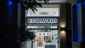 La entrada a EPIDAVROS, con fachada de vidrio, un mostrador de recepción y un interior moderno visible a través de las puertas.