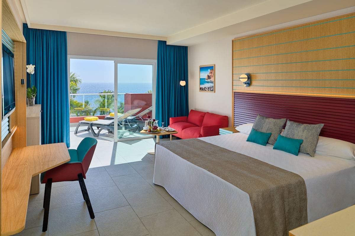 A hotel room with a bed, a desk, a chair, a sofa, and a balcony with a view of the sea.