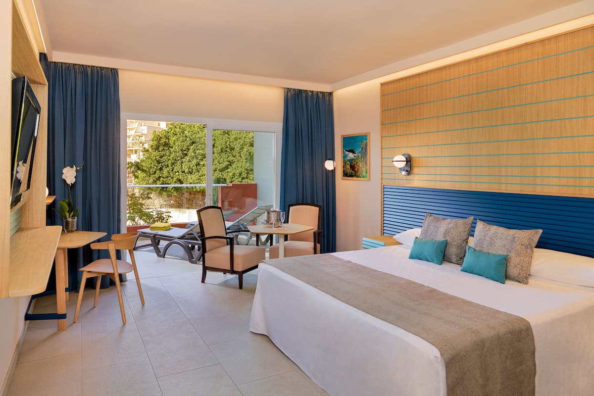 A hotel room with a bed, a desk, chairs, and a balcony with a view of greenery.