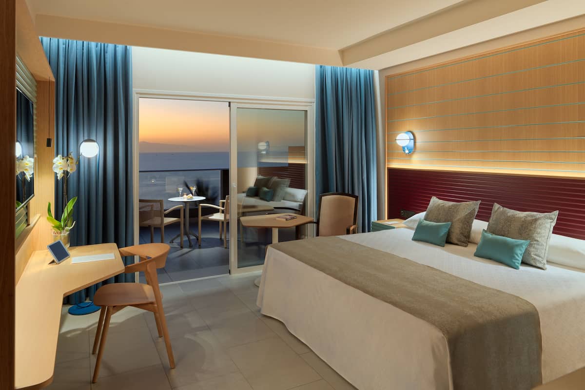 A hotel room with a bed, a desk with a chair, a small table, and a balcony with a view of the sunset.
