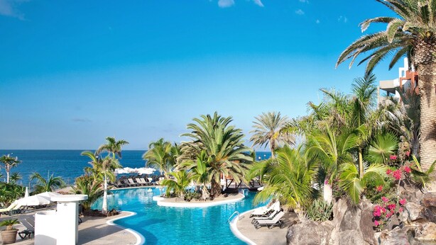 A resort pool with a view of the ocean, surrounded by palm trees and lounge chairs. - Roca Nivaria Gran Hotel (Adeje)