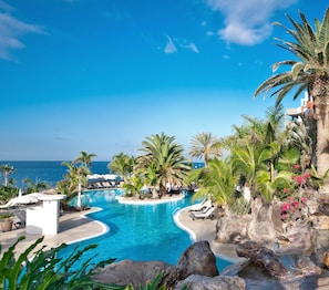 Zwembad van een resort met uitzicht op de oceaan, omringd door palmbomen en ligbedden.