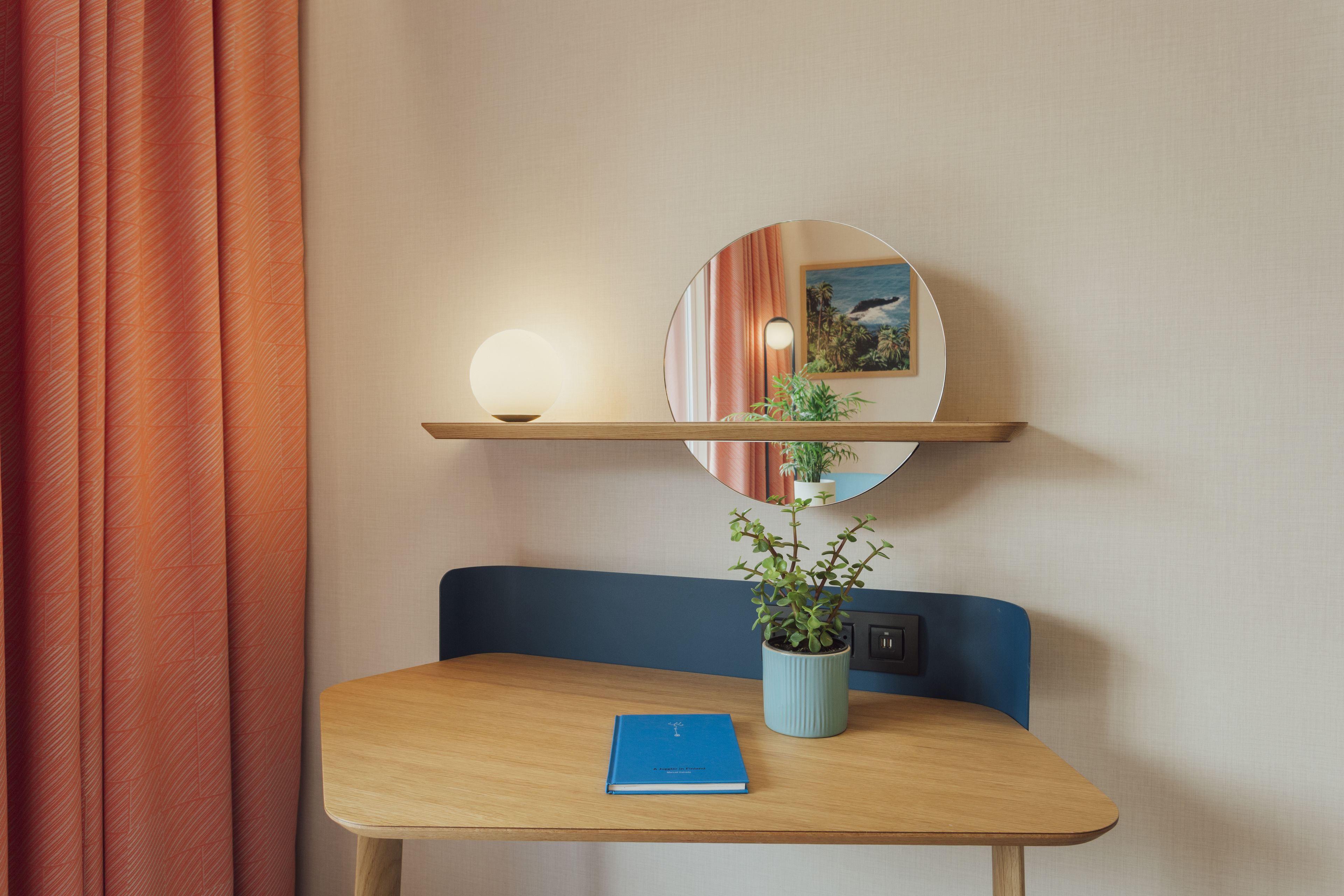 A room with a wooden table, a blue chair, a potted plant, a round mirror, and a wall-mounted shelf with a lamp and a framed picture.